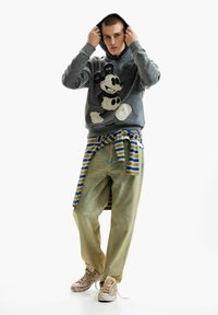 Fiatal férfi kapucnit igazít, aki szürke Mickey egér mintás kapucnis pulóvert visel, derékban megkötött csíkos inget, bő farmert és kopott tornacipőt fehér háttéren.