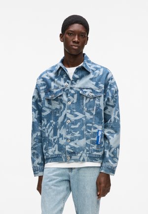 Noor mees kannab valget särk ja helesiniseid teksaseid ning sinist mustrilist teksajopet, seistes ühevärvilise valge tausta ees.