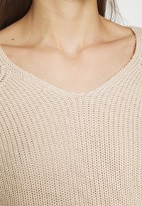 Beżowy sweter dziany z dekoltem w serek, otwartym splotem i prążkowanymi detalami na rękawach i korpusie.
