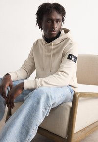 Felpa beige con cappuccio e lacci, caratterizzata da un'etichetta nera C.K. sul braccio, abbinata a jeans in denim azzurri chiari, seduta su una sedia di legno.