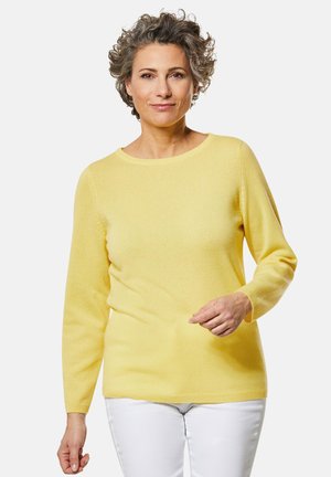 Pull en maille jaune à manches longues et col rond, présentant une texture lisse et une coupe décontractée. Porté avec un pantalon blanc.