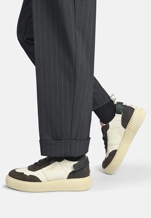 Person iført sorte bukser med nålestriber og hvide og brune sneakers med tykke beige såler, vist fra de nedre lægge og ned.