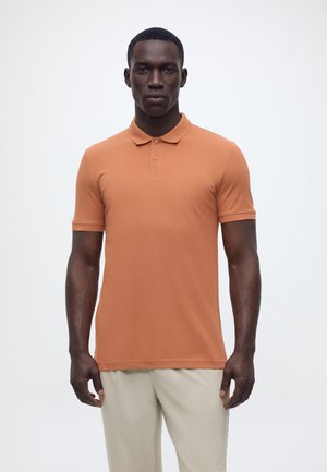 Mężczyzna stojący przodem, ubrany w pomarańczową krótką koszulkę polo i jasnobeżowe spodnie na jednolitym białym tle.