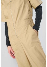 Combinaison beige en tissu léger avec des manches courtes, fermeture éclair à l'avant, deux poches latérales et des détails de couture subtils.