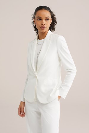 Ung kvinde med krøllet hår iført hvid blazer, hvide bukser, lagdelte halskæder og creoler, der kigger til siden med hænderne i lommerne.