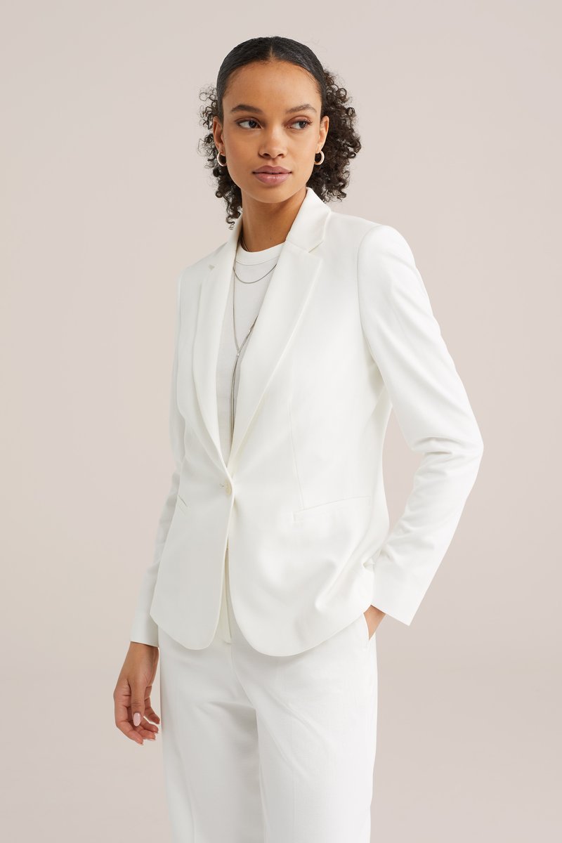 Mladá žena s kudrnatými vlasy, která má na sobě bílý blejzr, bílé kalhoty, vrstvené náhrdelníky a kruhové náušnice, dívá se stranou s rukama v kapsách.