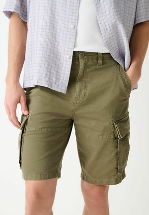 Muž nosiaci khaki krátke cargo nohavice s klopovými vreckami a svetlú károvanú košeľu s krátkym rukávom cez biele rebrované tielko.