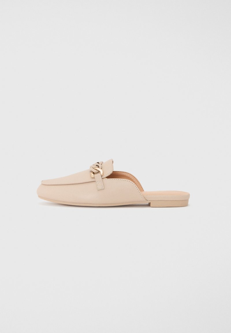 Chaussure à enfiler en cuir beige avec un petit talon, un bout arrondi et une garniture décorative dorée sur le dessus. Texture lisse et design minimaliste.