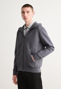 Sudadera gris con cremallera, fabricada en tela suave y de corte relajado. Presenta capucha con cordón, bolsillos laterales y un sutil logo en el pecho.