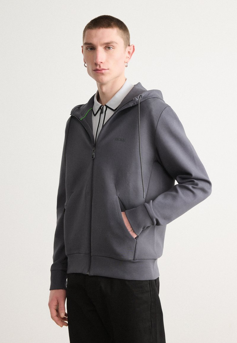 Sudadera gris con cremallera, fabricada en tela suave y de corte relajado. Presenta capucha con cordón, bolsillos laterales y un sutil logo en el pecho.
