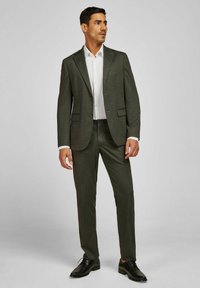 Dunkelgrüner, strukturierter Anzug mit maßgeschneiderter Passform, kombiniert mit einem weißen Hemd mit Knöpfen. Schwarze Schuhe vervollständigen den formellen Look.