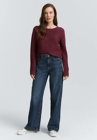 Bordowy prążkowany sweter z długimi rękawami w połączeniu z jasnoniebieskimi jeansami o szerokich nogawkach i białymi tenisówkami na modelce na neutralnym tle.