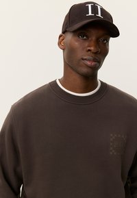 Σκούρο καφέ φούτερ με ριμπ λαιμόκοψη και διακριτικό λογότυπο. Συνδυασμένο με σκούρο καπέλο που φέρει λευκά κεντημένα γράμματα.