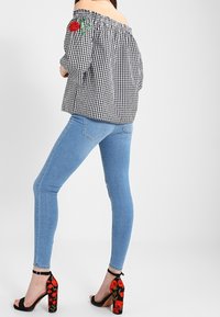 Top off-the-shoulder in gingham bianco e nero con ricami di rose verdi e rosse, abbinato a jeans skinny blu chiaro e sandali con tacco floreali.