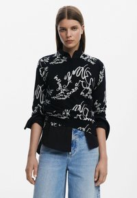 Svart skjorta med vitt abstrakt handskriftmönster, hög krage och rullade ärmar. Kombinerad med ljusblå denimjeans.