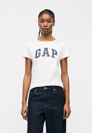 Mujer joven con el cabello trenzado, vistiendo una camiseta blanca con el logo de GAP y jeans oscuros de talle alto, de pie frente a un fondo liso.