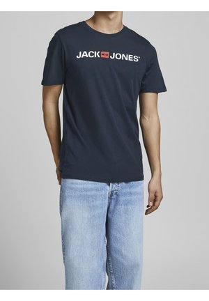 Mand iført en mørkeblå Jack Jones T-shirt og lyseblå jeans, stående mod en ensfarvet grå baggrund.