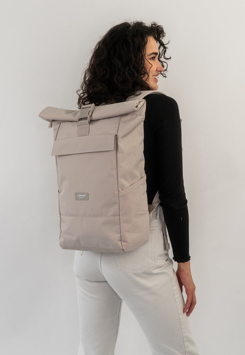 Sac à dos roulé gris clair de forme rectangulaire, fabriqué en tissu résistant, avec une poche avant à rabat et des bretelles ajustables.