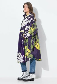 Kleurige bloemenregenjas in paars, groen en geel, met een capuchon, lange mouwen en zijzakken, gelaagd over blauwe jeans en metalen laarzen.