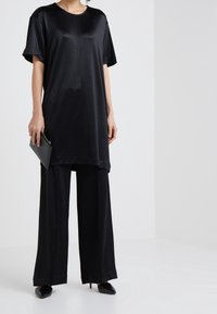 Svart, kortärmad tunika med lös passform, gjord av slät tyg, kombinerad med vida svarta byxor och spetsiga svarta skor, hållande en kuvertväska.