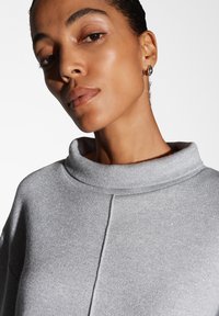 Top grigio texture con colletto alto e lieve luccichio, caratterizzato da una vestibilità rilassata e dettagli delle cuciture lungo la parte anteriore.