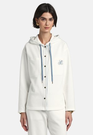 Žena oblečená v bielej mikine na gombíky s modrými šnúrkami a vyšívaným "L" na ľavej náprsnej vrecku, v kombinácii s bielymi nohavicami rovnakej farby.