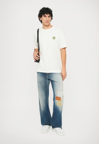 Weißes T-Shirt mit einem gelben Smiley-Patch, kombiniert mit blauen, verwaschenen weiten Jeans mit einem Patchwork-Design. Weiße Sneaker, schwarzer Riemen der Tasche.