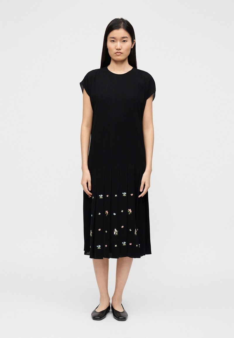 Robe noire plissée avec manches courtes, ornée de broderies florales colorées sur l'ourlet. Tissu lisse avec une silhouette fluide.