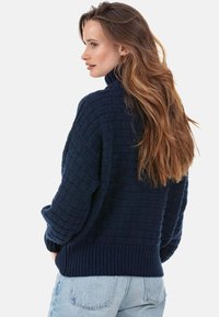 Pull en tricot bleu marine avec un motif texturé, col montant et poignets côtelés, associé à un jean en denim bleu clair.