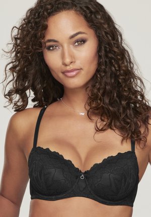 Frau mit lockigem Haar, die einen schwarzen Spitzen-BH und eine silberne Halskette mit dem Wort "LOVE" trägt, vor neutralem Hintergrund.