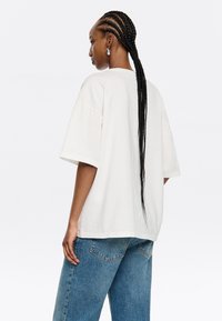 Camiseta blanca de gran tamaño, fabricada en algodón, con mangas cortas y cuello redondo, combinada con jeans de denim azul claro, de corte relajado, con costura trasera visible.