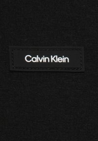 Musta kangas, jossa on tekstuuripinta, sisältää suorakulmaisen mustan etiketin, jossa on valkoisella tekstillä teksti "Calvin Klein."
