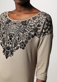 Beige top featuring black floral embroidery along the neckline and shoulders, with a scoop neckline and three-quarter sleeves.