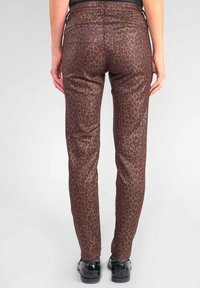 Pantalons imprimés léopard, marron et beige, avec une coupe ajustée, un éclat subtil et des poches arrière. Portés avec des chaussures noires.