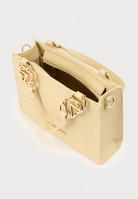 Κίτρινη τσάντα ώμου από υφή δέρματος με δύο ακροδέκτες σε χρυσό τόνο, κλείσιμο με φερμουάρ και διπλές λαβές. Το εσωτερικό εμφανίζει καφέ φόδρα.