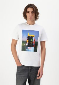 White cotton t-shirt featuring a graphic print of a backpack with yellow accents and rubber boots, set against a blue sky background.