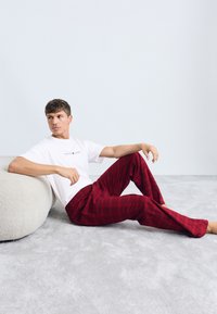 T-shirt blanche avec logo, pantalon écossais rouge, coupe décontractée. Texture douce, design minimal, assis sur un coussin rond en molleton gris clair.