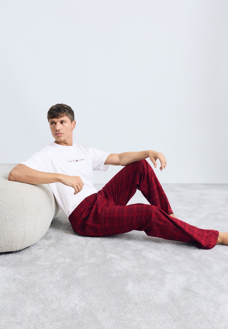 T-shirt blanche avec logo, pantalon écossais rouge, coupe décontractée. Texture douce, design minimal, assis sur un coussin rond en molleton gris clair.