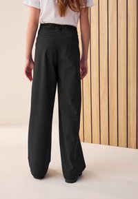 Pantalon noir à jambes larges avec taille haute, poches arrière et texture lisse. Le modèle est montré de dos devant un mur en bois.