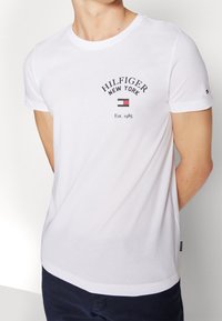 Camiseta de algodón blanca de manga corta, con escote redondo, que presenta el texto "HILFIGER NEW YORK" y un pequeño logotipo de bandera impresos en el pecho.