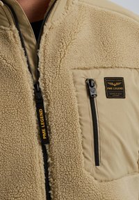 Béžová bunda s chlpatým, textúrovaným fleece predným dielom a hladkým zadným dielom. Obsahuje zipsovú náprsníkovú kapsu a nášivku značky.