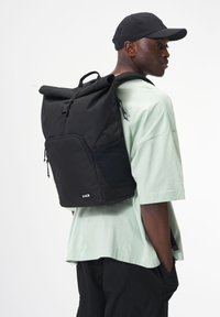 Schwarzer Rolltop-Rucksack mit verstellbaren Trägern, Fronttasche und seitlichem Mesh-Einsatz, kombiniert mit einem mintgrünen Oversize-Shirt.