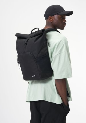 Black roll-top backpack featuring adjustable straps, front pocket, and mesh side panel, paired with a mint green oversized shirt.