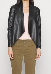 Veste cropped en simili cuir noir avec un devant drapé, associée à un top rose clair à côtes et un pantalon vert olive. Texture lisse et coupe ajustée.