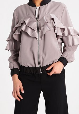 Personne portant une veste zippée violet clair avec des volants sur la poitrine et les manches, associée à un pantalon noir.