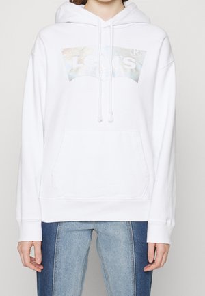 Biała bluza z kapturem Levi's z metalicznym logo na przodzie, kieszenią kangurka i ściągaczem przy kapturze, noszona z dwutonowymi niebieskimi dżinsami.