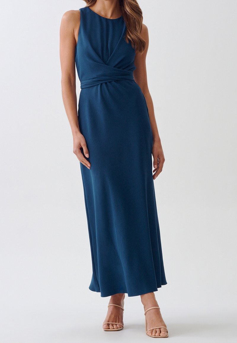 Femme portant une robe bleu marine sans manches, longue jusqu'au sol, avec un détail torsadé à la taille, assortie à des talons beiges à lanières.