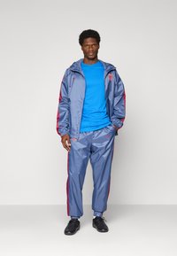 Mann steht da und trägt eine blaue Sportjacke und Hose mit roten Streifen, ein blaues Hemd und schwarze Sportschuhe vor einem einfarbigen Hintergrund.