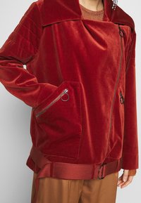 Mujer con chaqueta de terciopelo roja con cremallera y tiradores en forma de anillo plateado, con cuello texturizado, sobre un suéter y pantalones marrones.