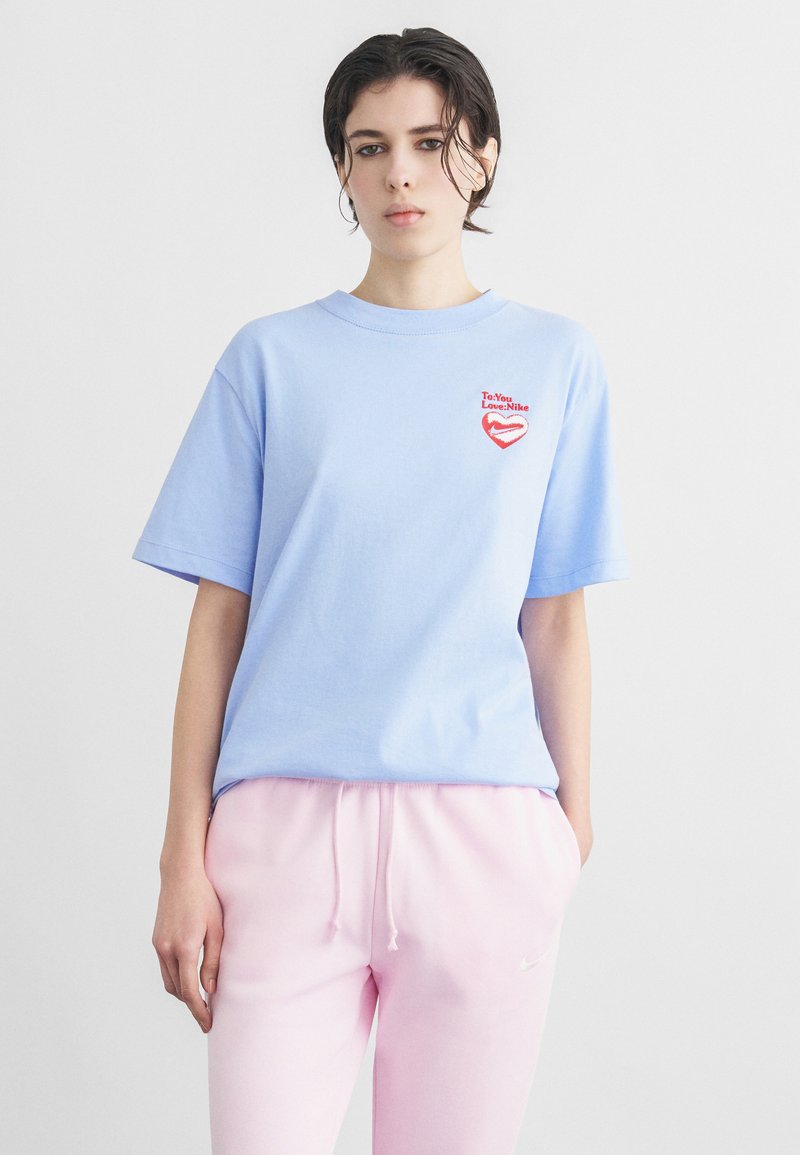 Άτομο που φοράει ένα ανοιχτό μπλε T-shirt της Nike με κόκκινη καρδιά λογότυπο και ροζ φόρμα Nike, στέκεται μπροστά σε ένα απλό φωτεινό φόντο.